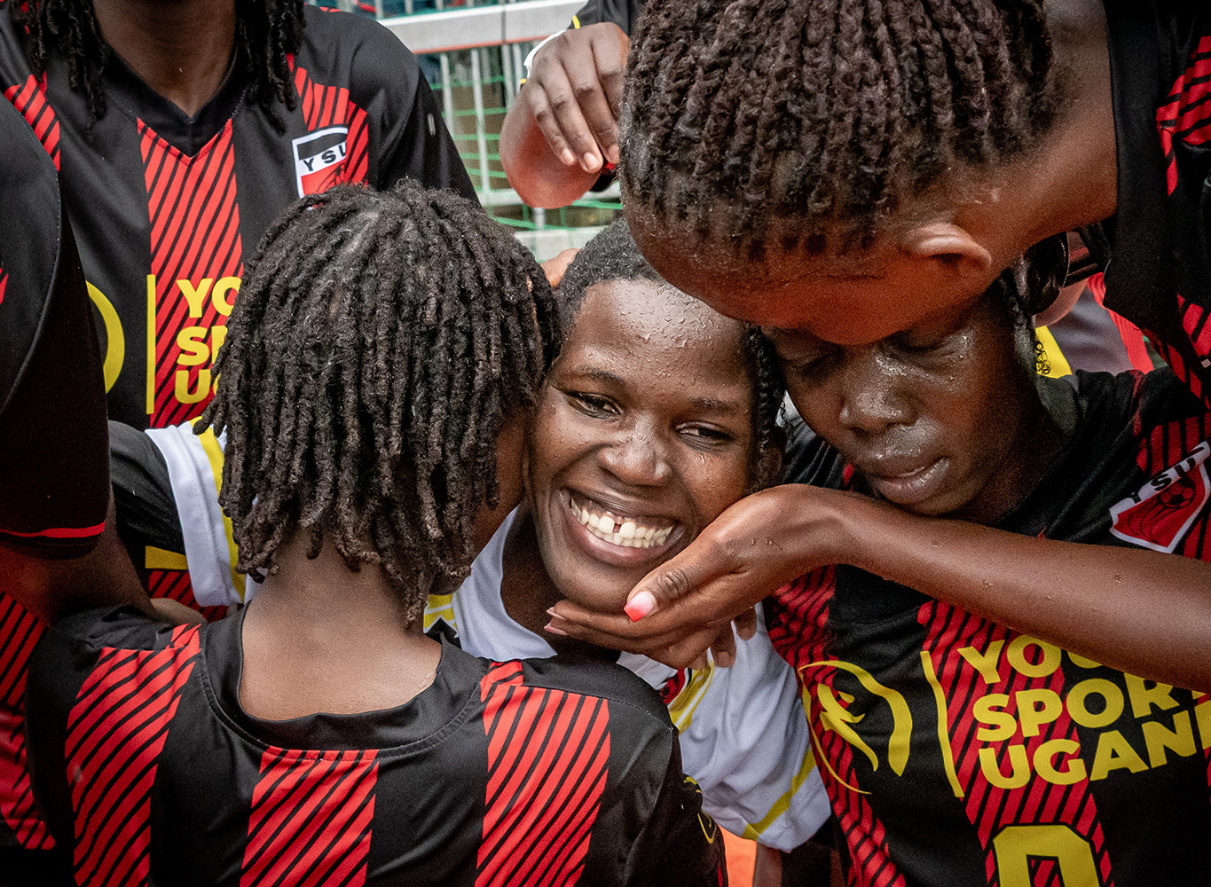Smiling football player surrounded by teammates in black and red jerseys during a close team huddle on the pitch.